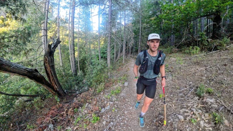 A trail runner wearing a cap and hydration vest jogs along a narrow, rocky path through a dense forest with tall trees and greenery.